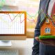 Photo of man holding a house next to a computer with stock charts